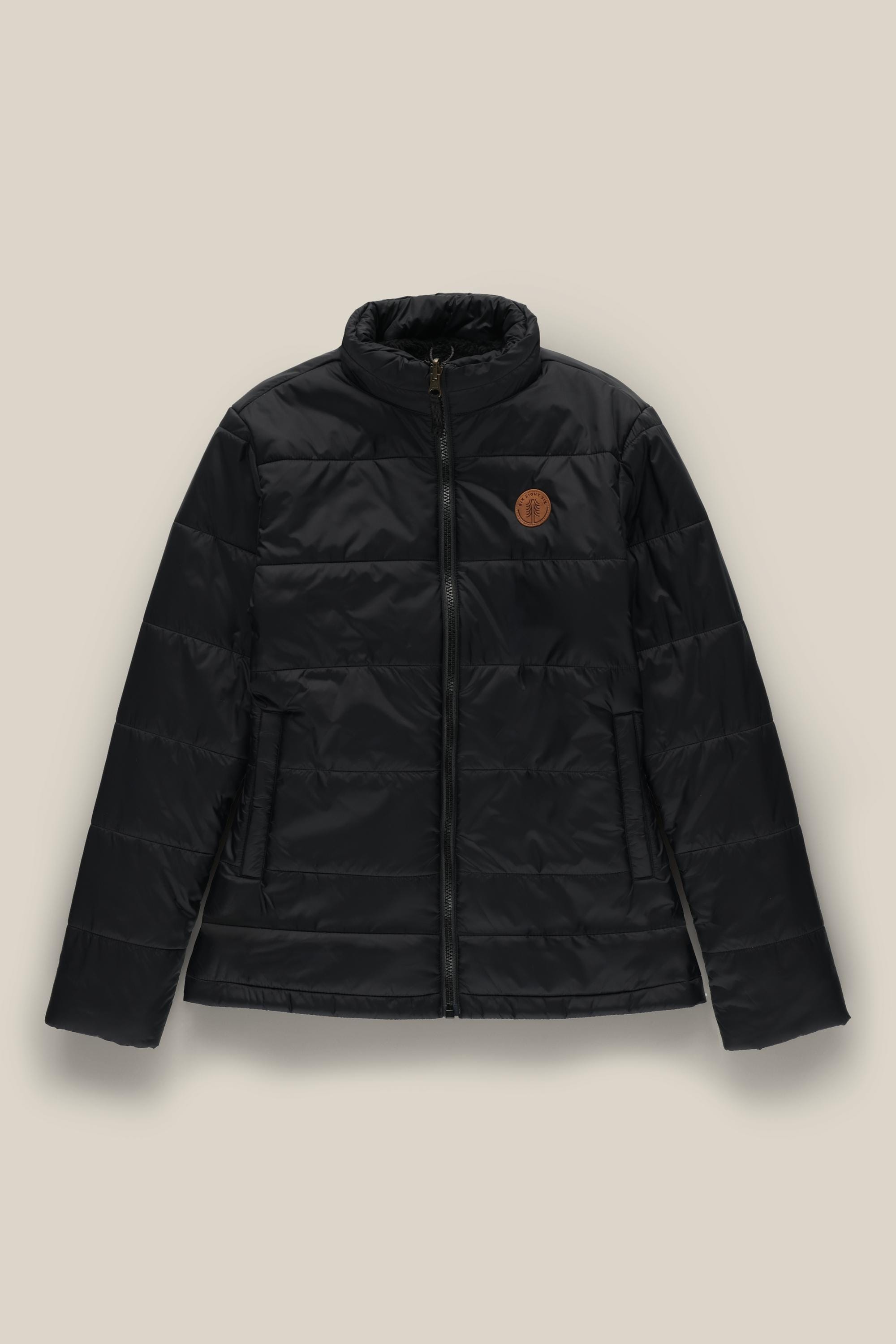 The 686 Womens Thermal Puff Insulated Jacket by 686 is a black quilted polyester jacket featuring a front zipper, two side pockets, and a brown circular patch on the left chest, shown against a beige background.