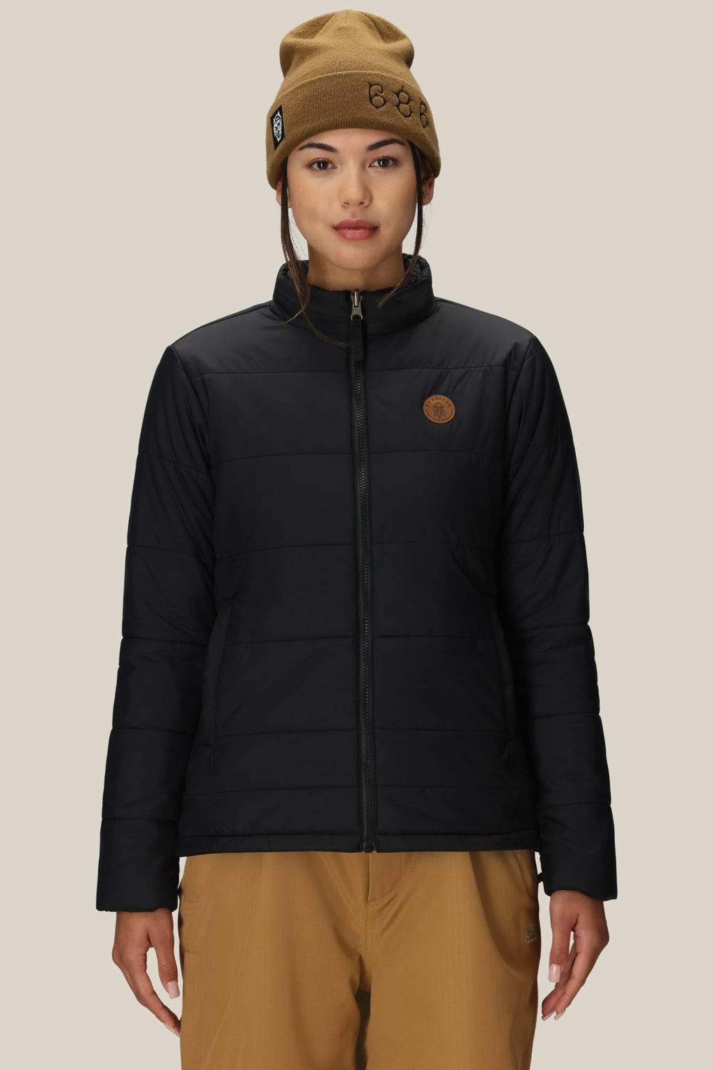 A person wearing a brown beanie, tan pants, and the 686 Womens Thermal Puff Insulated Jacket by 686 with a circular chest patch stands facing forward against a plain beige background.