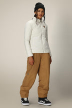 A woman in a 686 Womens Thermal Puff Insulated Jacket, tan baggy pants, black beanie, and black Vans sneakers stands against a neutral background, looking at the camera with a slight smile.