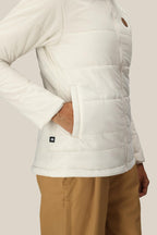 A person wearing the 686 Womens Thermal Puff Insulated Jacket with their right hand in a side pocket, paired with tan pants, stands against a plain beige background.