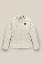 The 686 Womens Thermal Puff Insulated Jacket by 686 is a polyester puffer with a front zipper, side pockets, high collar, and a round brown patch on the left chest. Shown against a plain beige background.