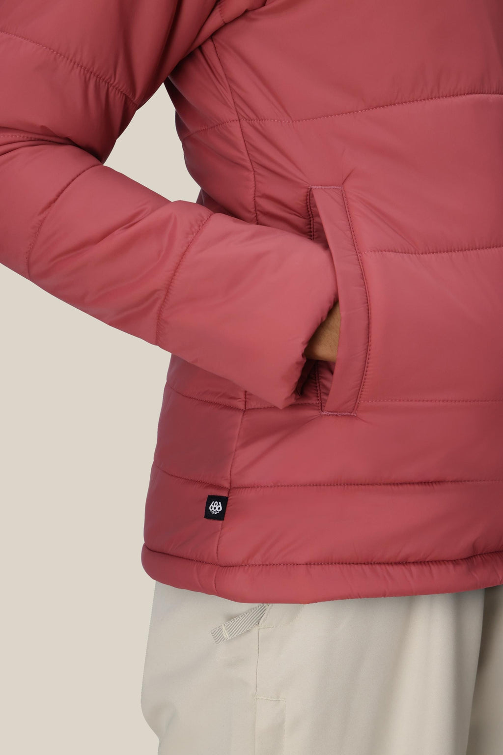 A person wearing the 686 Womens Thermal Puff Insulated Jacket in mauve, with one hand in the pocket and light-colored pants, stands against a plain light background.