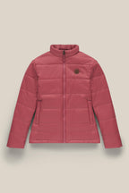 The 686 Womens Thermal Puff Insulated Jacket by 686 features a pink quilted design, high collar, front zipper, two side pockets, a circular brown chest patch, and is made from soft polyester. Shown against a light beige background.
