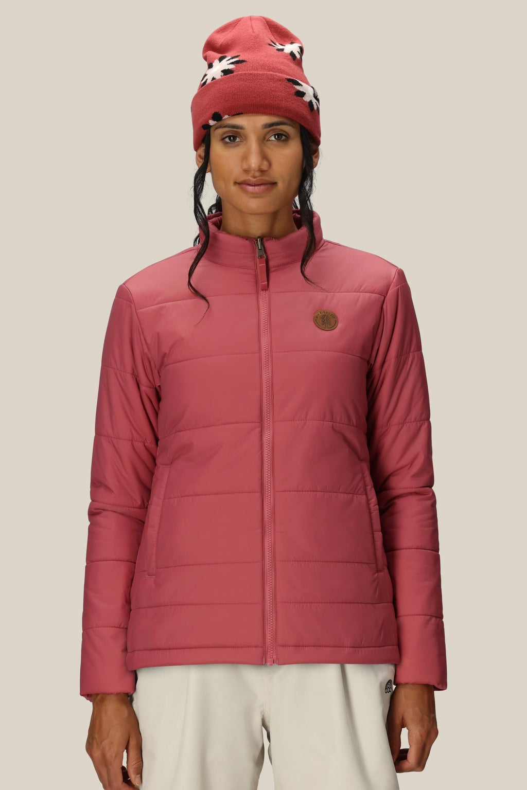 A woman in the 686 Womens Thermal Puff Insulated Jacket and white pants, wearing a red beanie with white patterns, stands facing forward against a plain beige background.