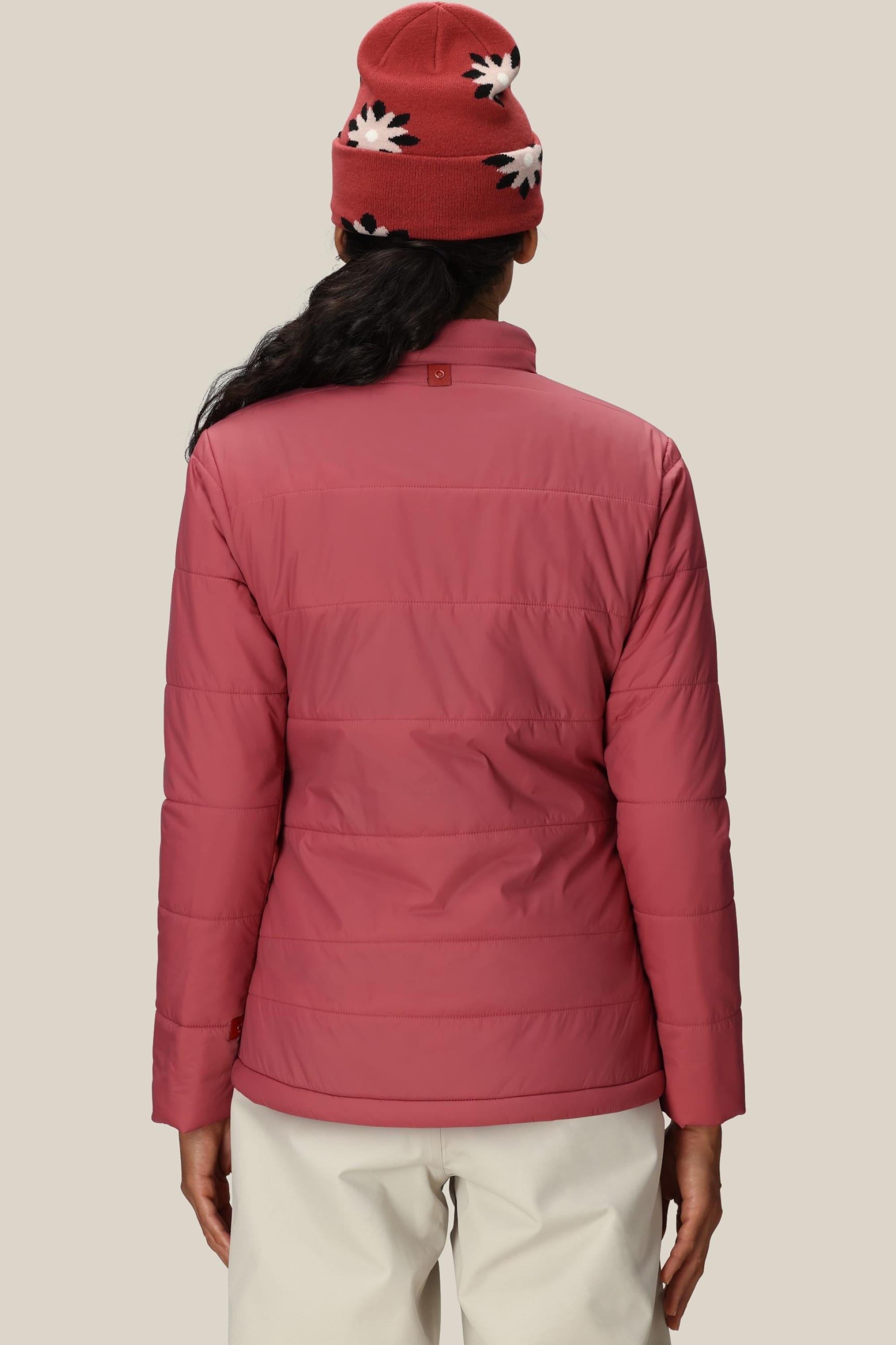 A person faces away, wearing the 686 Womens Thermal Puff Insulated Jacket in pink and white pants, with a red knit hat featuring white and black patterns, standing against a plain beige background.