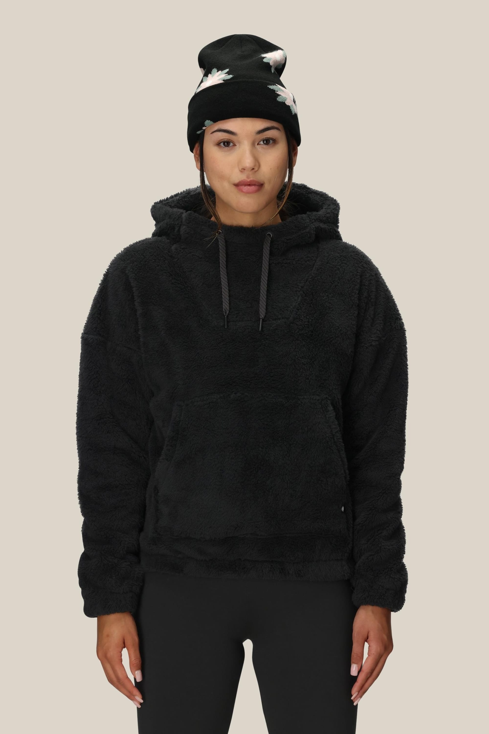 A person stands facing forward on a light background, wearing the 686 Womens Sherpa Hoody in black, paired with black leggings and a black beanie featuring white floral designs.