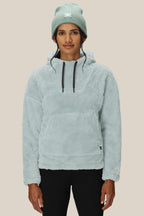 A woman in the 686 Womens Sherpa Hoody by 686, a light blue polyester jacket with a front pocket and matching beanie, stands against a plain beige background, facing forward with a relaxed expression.