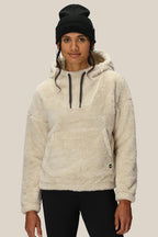 A woman in a black beanie, black leggings, and the 686 Women’s Sherpa Hoody—a cream-colored polyester jacket with a front pocket—stands facing forward against a plain light background.