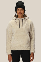 A woman wearing the 686 Womens Sherpa Hoody—a cream-colored, fuzzy polyester jacket with black drawstrings—stands facing forward against a plain beige background, paired with a black beanie.