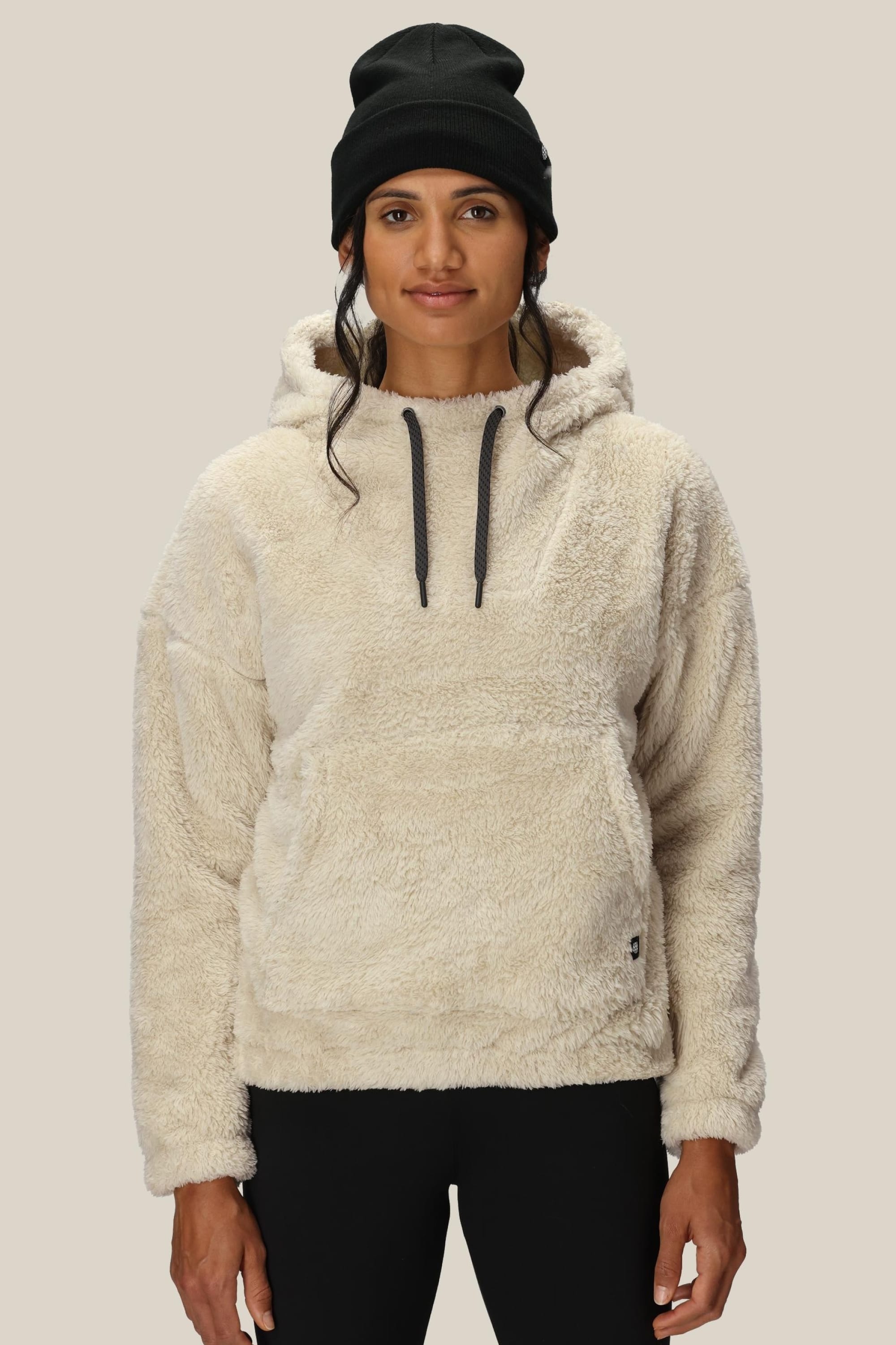A woman wearing the 686 Womens Sherpa Hoody—a cream-colored, fuzzy polyester jacket with black drawstrings—stands facing forward against a plain beige background, paired with a black beanie.
