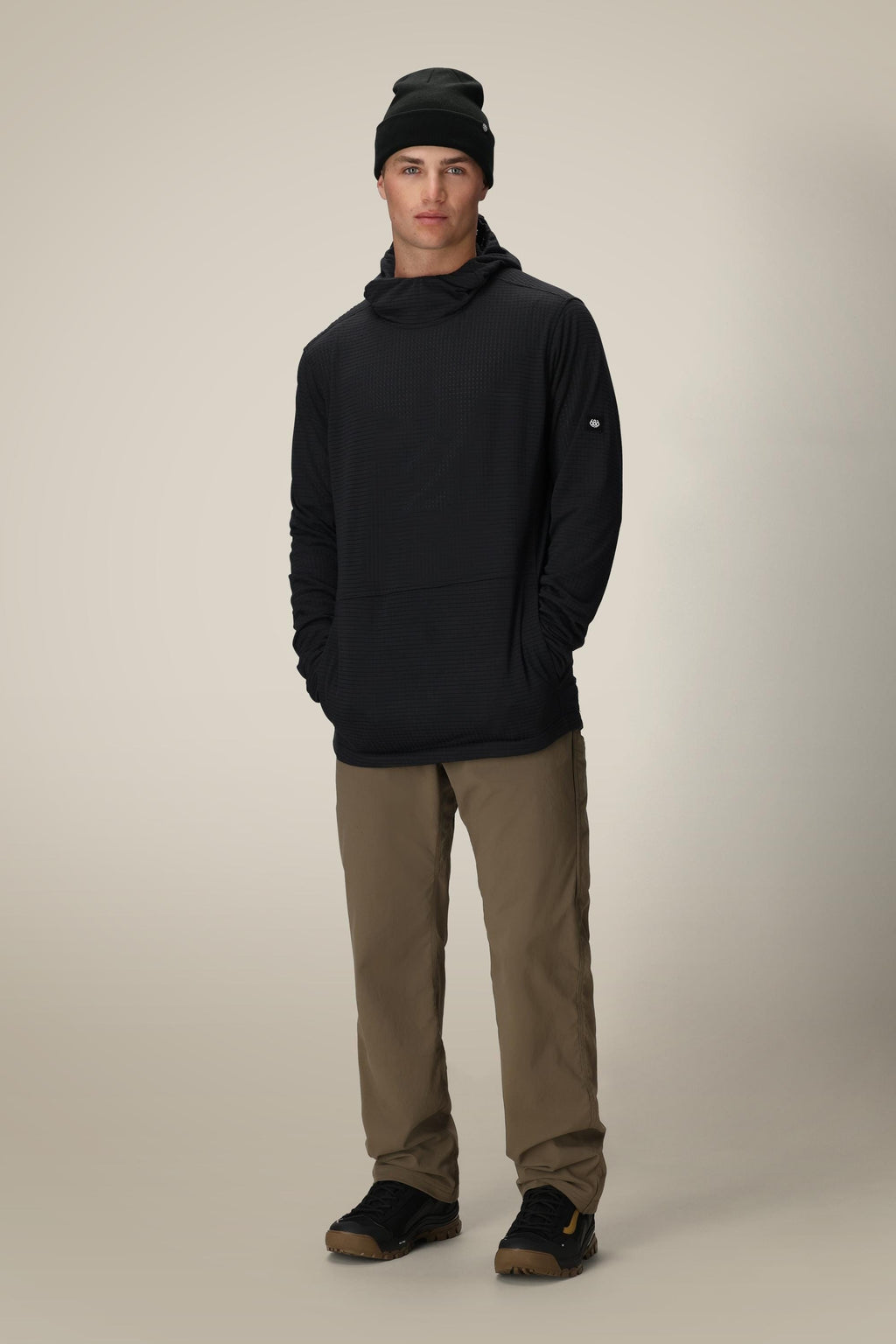 A person stands facing forward, hands casually in their pockets, wearing the 686 Men's Mission Grid™ Fleece Hoody in black with khaki pants and black shoes against a plain light background.