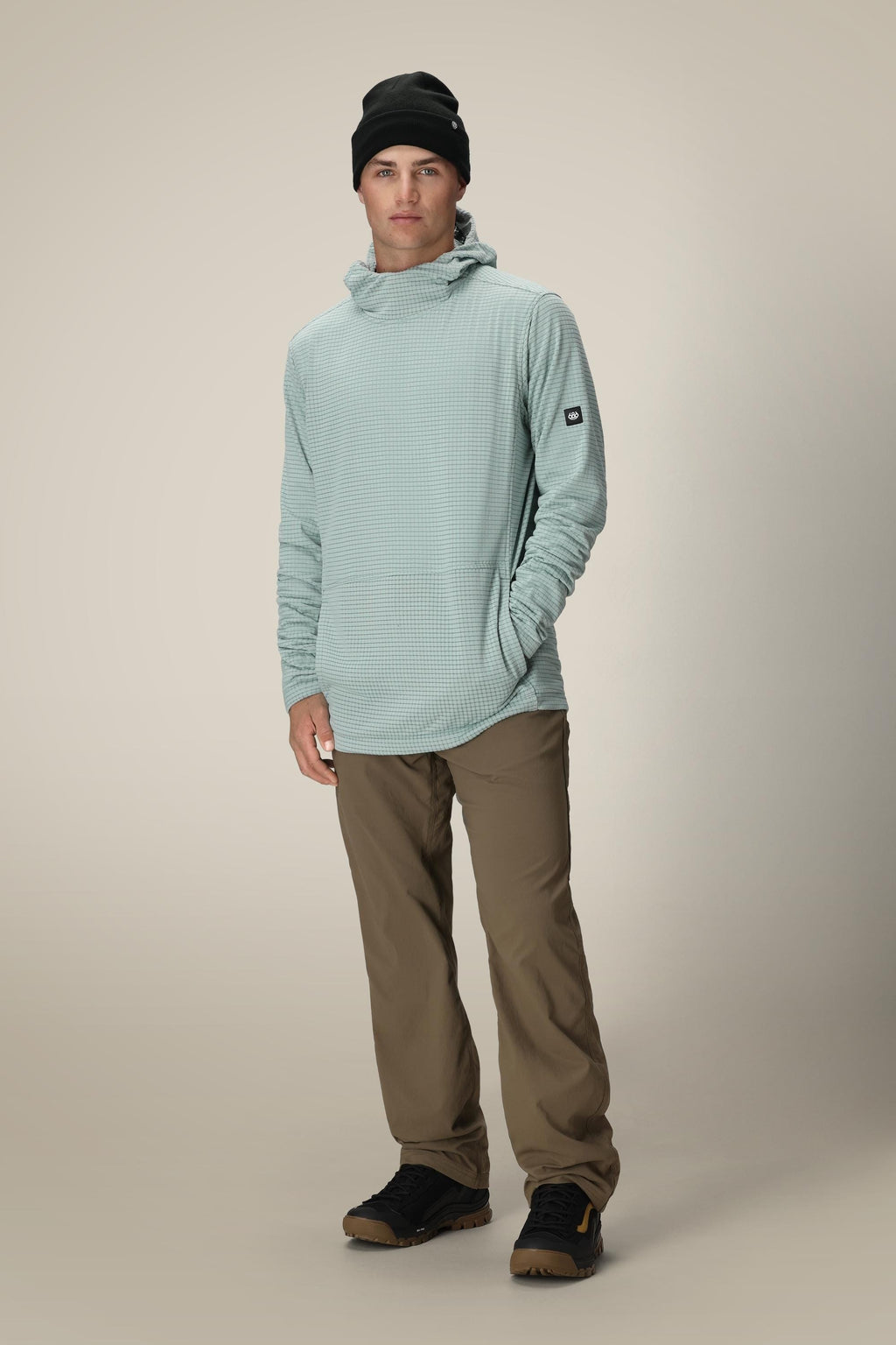 A young man wearing a black beanie, 686 Men's Mission Grid™ Fleece Hoody in light blue, khaki pants, and black sneakers stands facing the camera with his left hand in his pocket against a plain beige background.