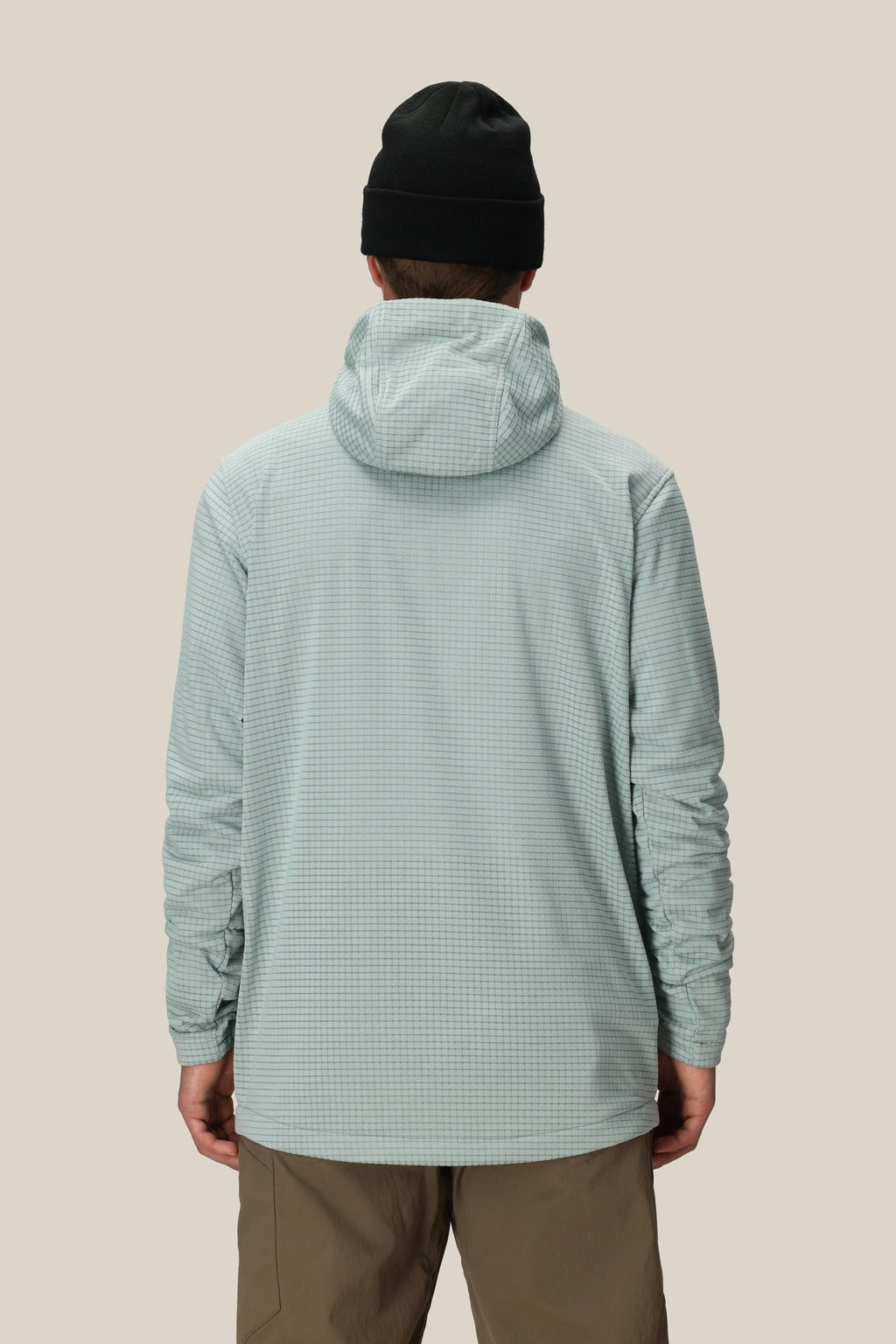 A person wearing a black beanie, khaki pants, and the 686 Men's Mission Grid™ Fleece Hoody by 686 in light blue stands with their back to the camera against a plain beige background.