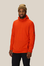 A man stands against a plain beige background, looking at the camera with a neutral expression. He is wearing a tan beanie, black pants, and the 686 Men's Mission Grid™ Fleece Hoody by 686 in orange.