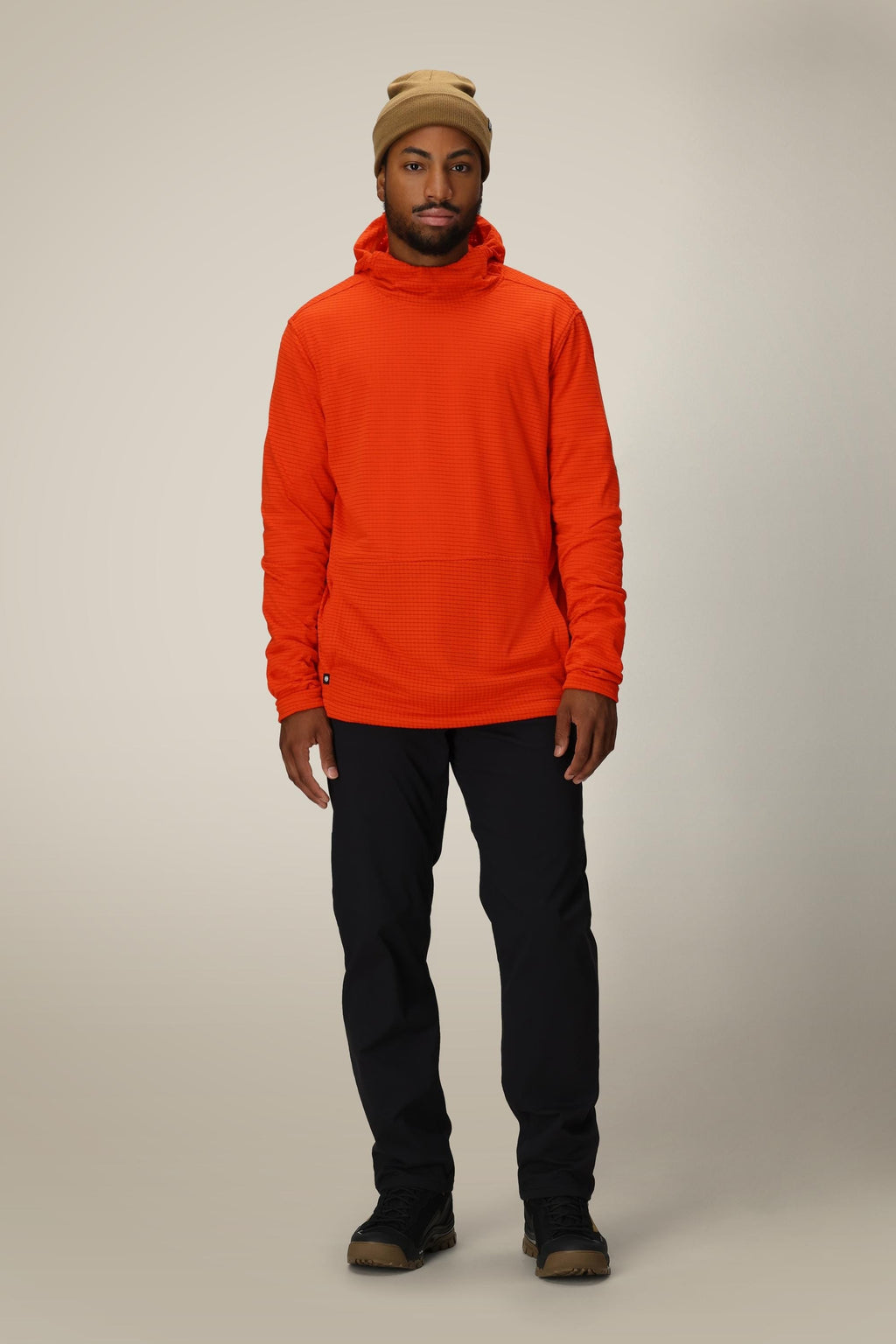 A man wearing a tan beanie, black pants, brown shoes, and a 686 Men's Mission Grid™ Fleece Hoody by 686 stands facing forward against a plain, light gray background.
