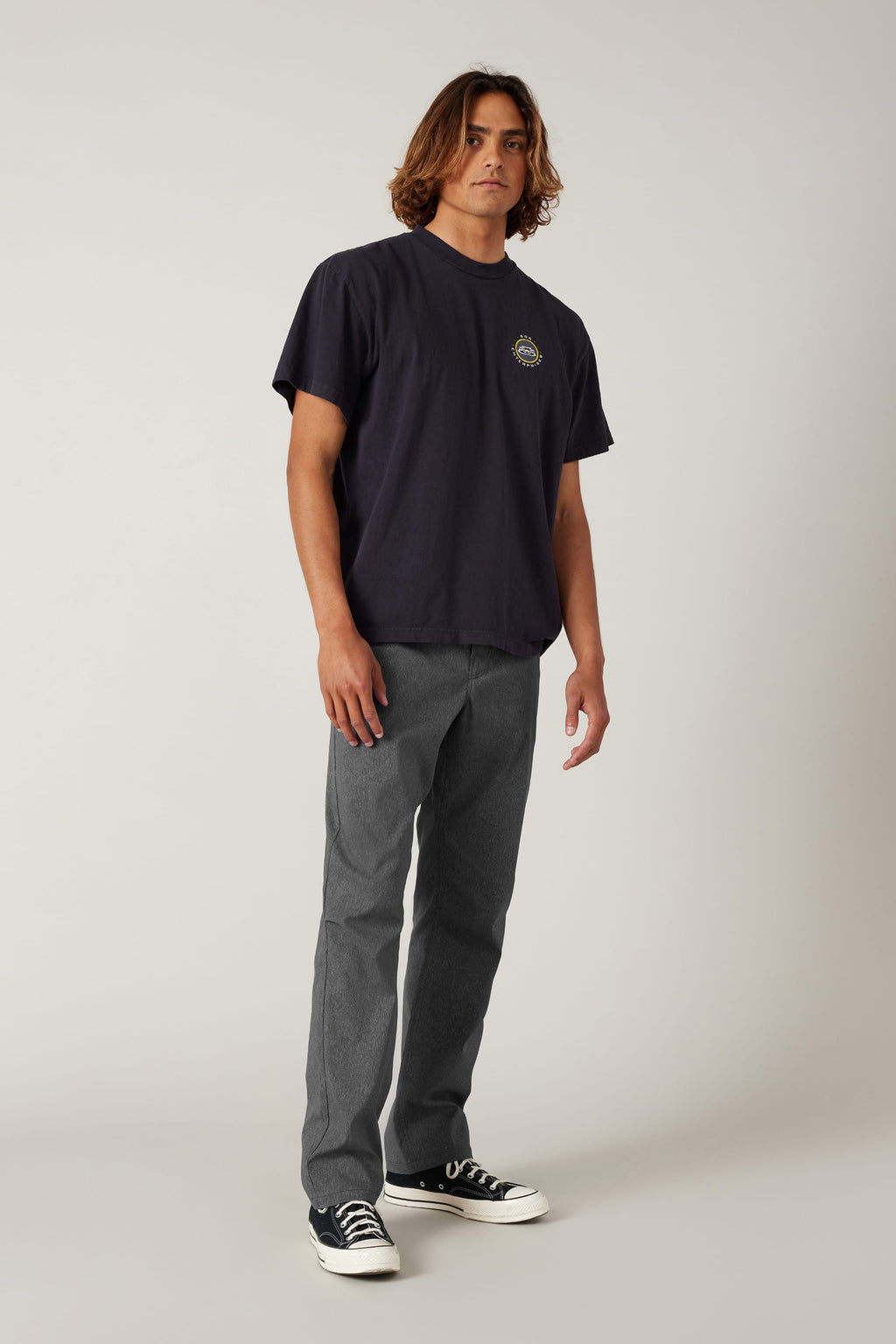 A young man with medium-length brown hair wears a loose black t-shirt, 686 Men's Everywhere® Pant - Relaxed Fit by 686 in gray, and black-and-white sneakers, standing against a plain light background.